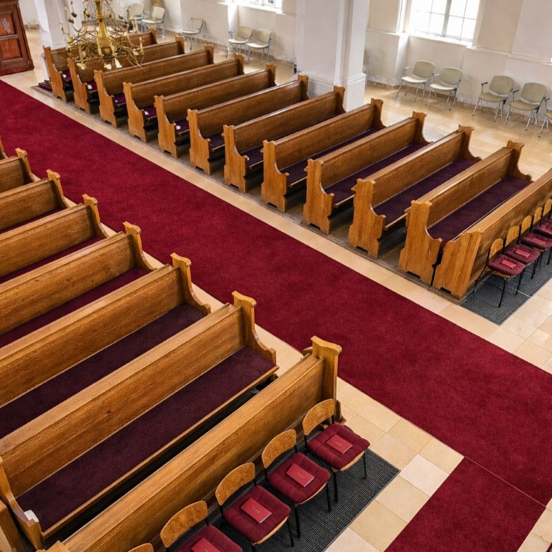 Innenraum einer Kirche mit Holz-Kirchenbänken und Stühlen, die bordeauxrote Filzauflagen haben.
